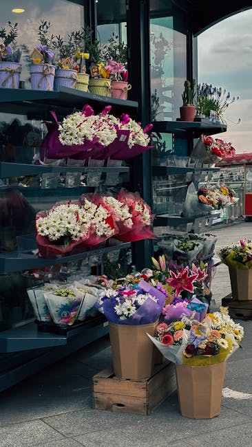 An outdoor display of fresh floral arrangements, including bouquets of white and pink chrysanthemums wrapped in pink paper, and various other flower bunches in purple, yellow, and mixed colors, arrang