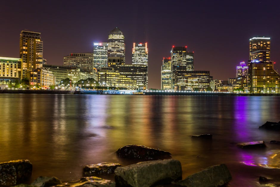 A clear night view of London's Canary Wharf skyline with illuminated modern skyscrapers reflecting vibrant lights on the calm river. In the foreground, wet rocks and stones are visible along the river